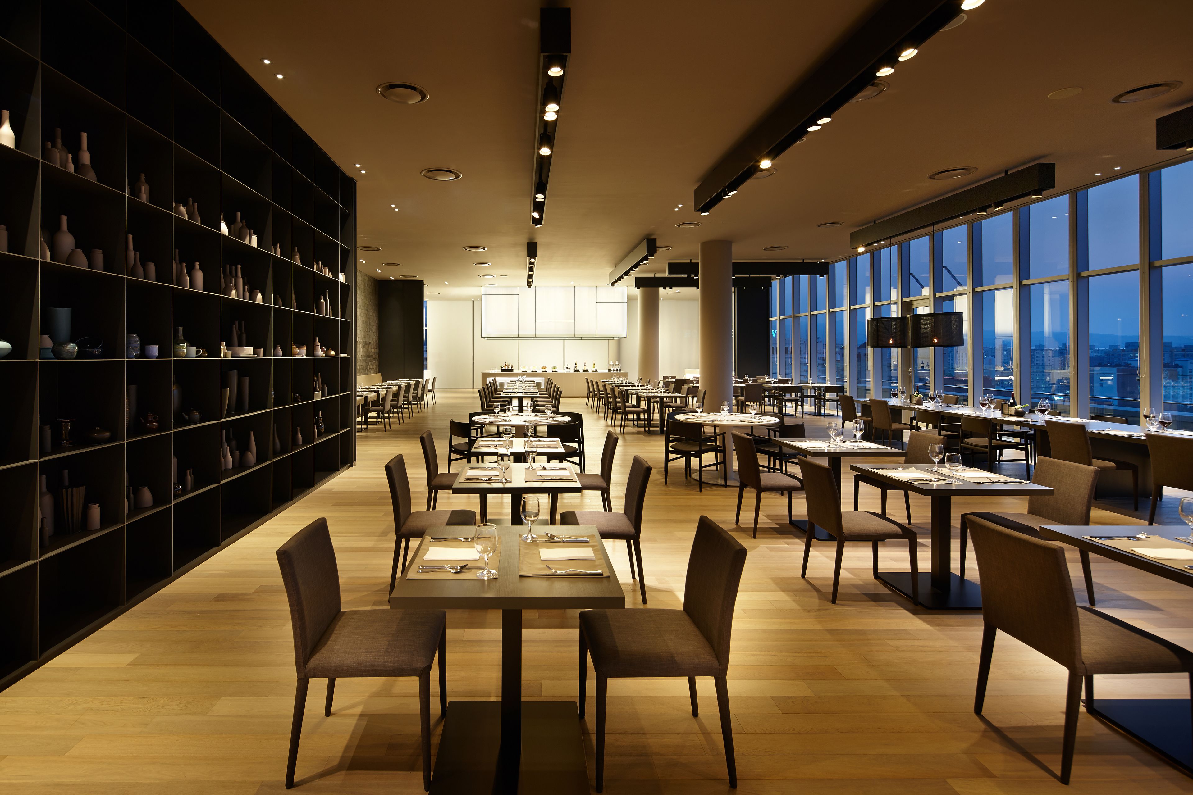 Um restaurante espaçoso com várias mesas e cadeiras, uma grande estante repleta de livros e objetos decorativos, e amplas janelas com vista para a cidade.