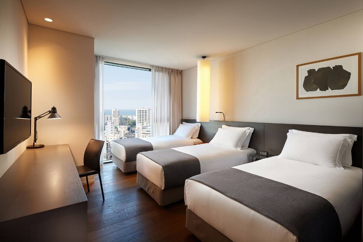 Habitación de hotel con dos camas, un escritorio, una silla, un televisor y un ventanal con vistas a la ciudad.
