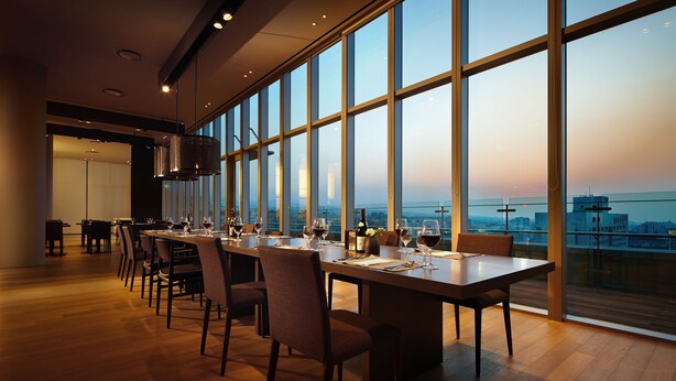 A spacious dining area with a long table set for a meal, large windows offering a city view, and modern interior design. - Shilla Stay Jeju Airport (Jeju City)