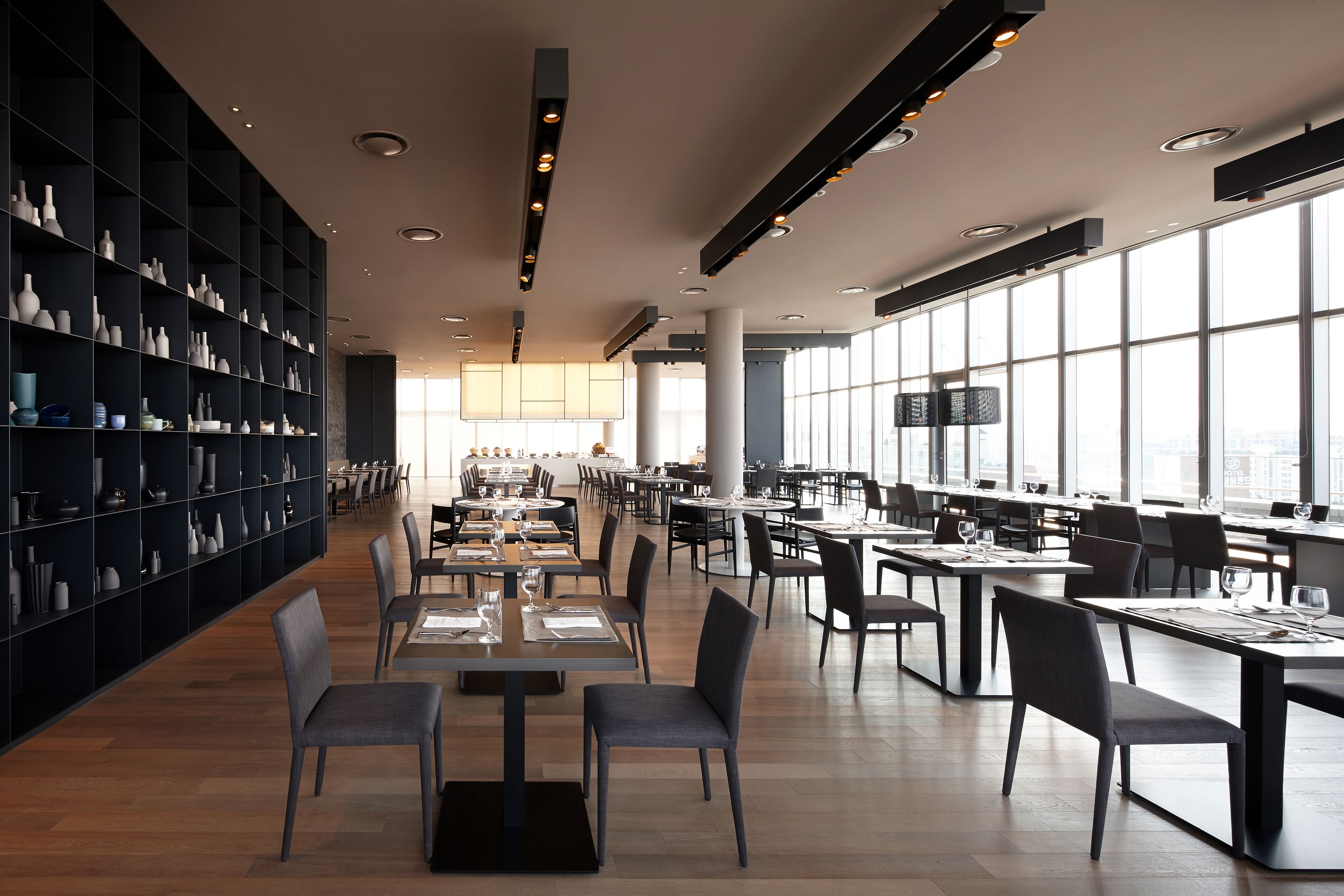 A modern dining area with tables and chairs, a large bookshelf, and large windows offering a city view.