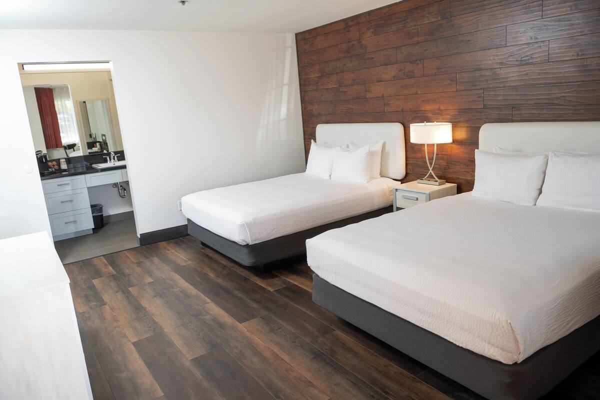 A hotel room with two beds, a wooden headboard, a nightstand with a lamp, a bathroom with a mirror, and a wooden floor.