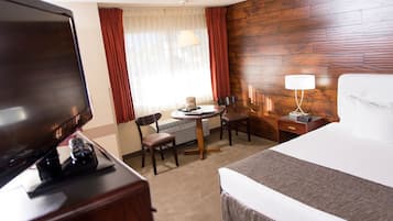 A hotel room with a bed, a TV, a desk with a chair, a lamp, and a window with curtains.