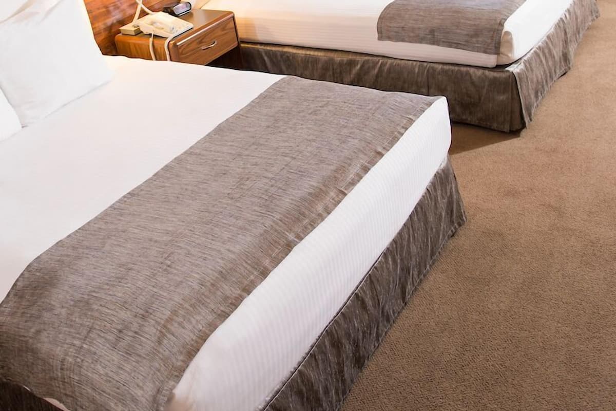 A hotel room with two beds, a wooden headboard, a small table with chairs, and a window with curtains.