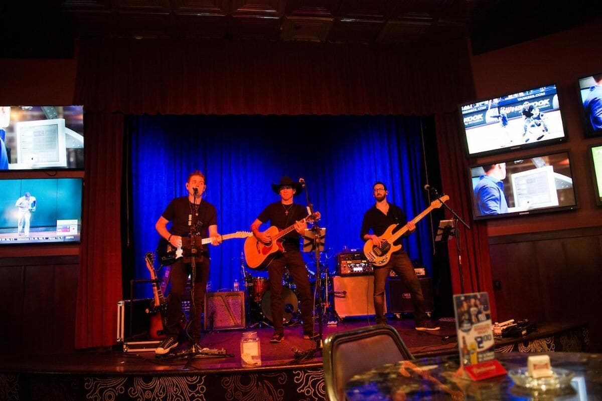 Een band treedt op in een theater met een blauwe gordijnenachtergrond, omringd door monitoren en een bar.