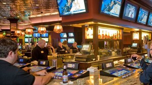 Un bar de casino con varias pantallas mostrando eventos deportivos, una zona de bar con botellas y clientes jugando a las máquinas tragamonedas.