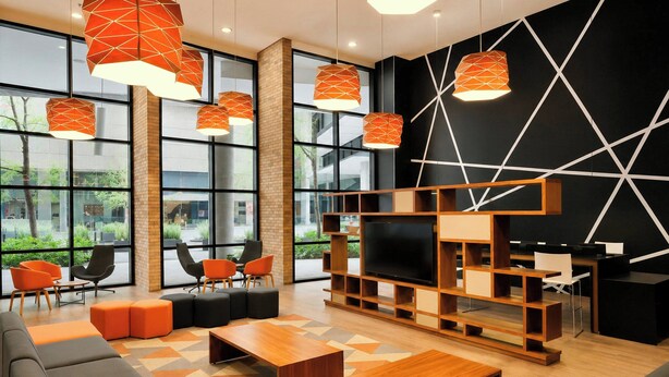 A modern lounge area with sofas, a coffee table, and a TV screen, featuring geometric wall patterns and large windows. - City Express Plus by Marriott Monterrey Nuevo Sur (Monterrey)