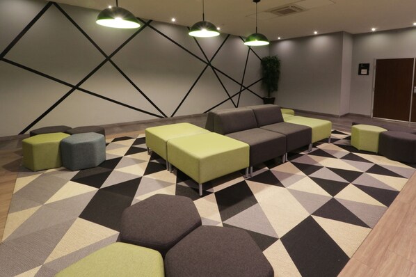 Un área de lounge moderna con alfombra de estampado geométrico, diversas opciones de asientos que incluyen sofás y otomanas, e iluminación colgante.