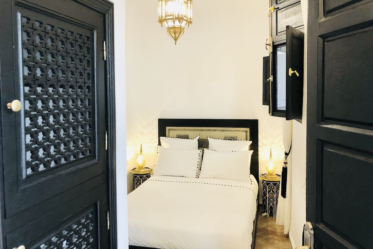 Une chambre à coucher comprenant un lit, deux tables de chevet avec des lampes, une décoration murale et une porte noire ornée d’un motif en treillis.