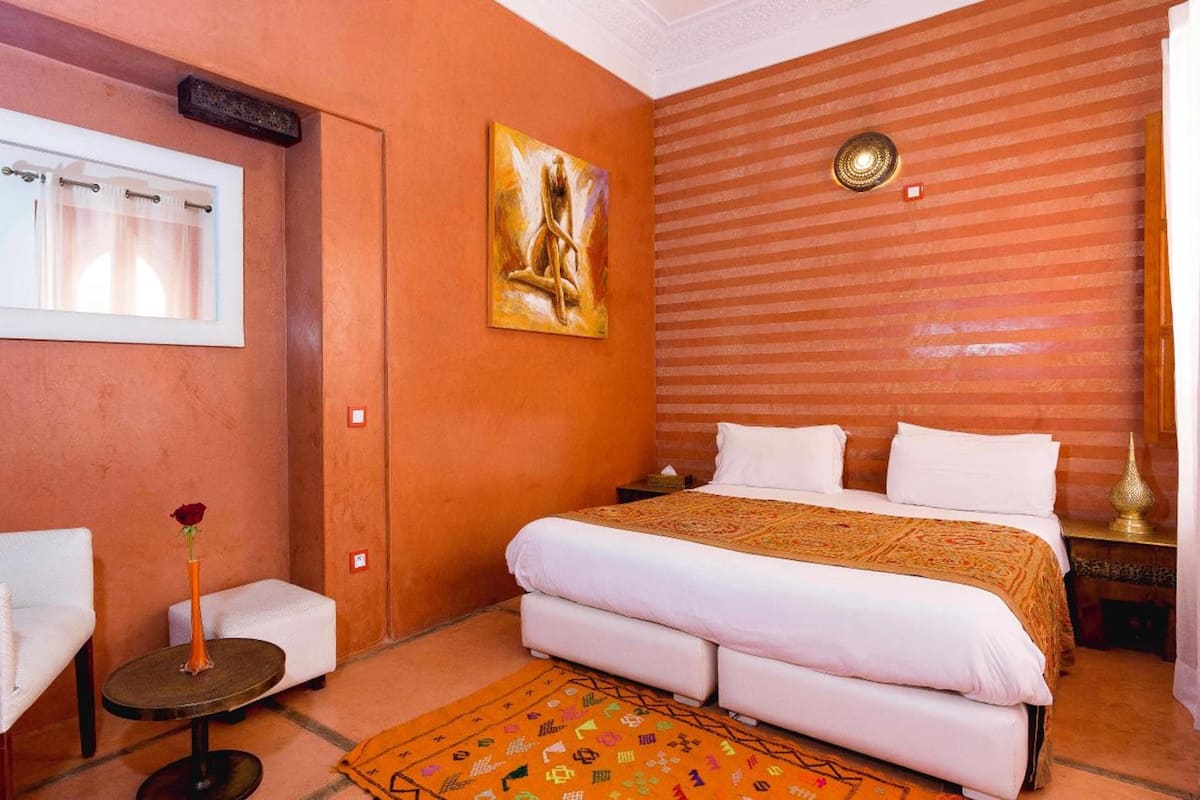 Une chambre d’hôtel avec un lit, une petite table, une chaise, un tableau et une fenêtre avec des rideaux.