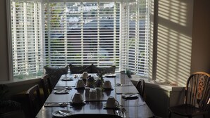 A dining area with a table set for four, large windows with blinds, and a view of a residential area. - South View Guest House (Lynton)