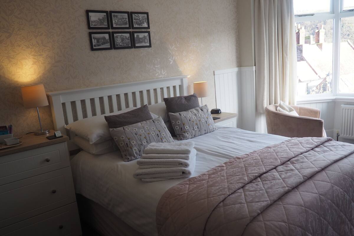 Ein ordentlich bezogenes Bett mit Kissen und Decke, ein Nachttisch mit Lampe, ein Stuhl und ein Fenster mit Blick auf Gebäude.