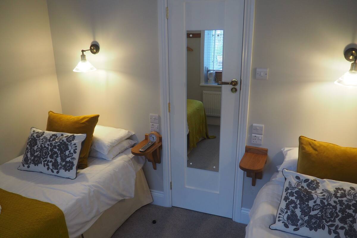 Ein Zimmer mit zwei Betten, jedes mit einem Kissen und einem Nachttisch mit einer Uhr. Eine Tür, ein Spiegel der ein Fenster reflektiert und eine Heizung.