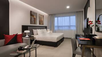 A modern hotel room with a large bed, a desk, a sofa, and a glass coffee table.