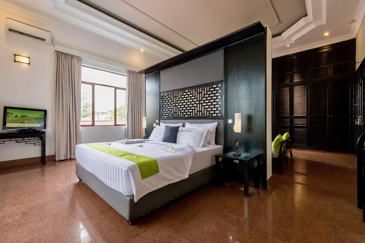 A hotel room with a large bed, a flat-screen TV, a desk with a computer, and a window with curtains.