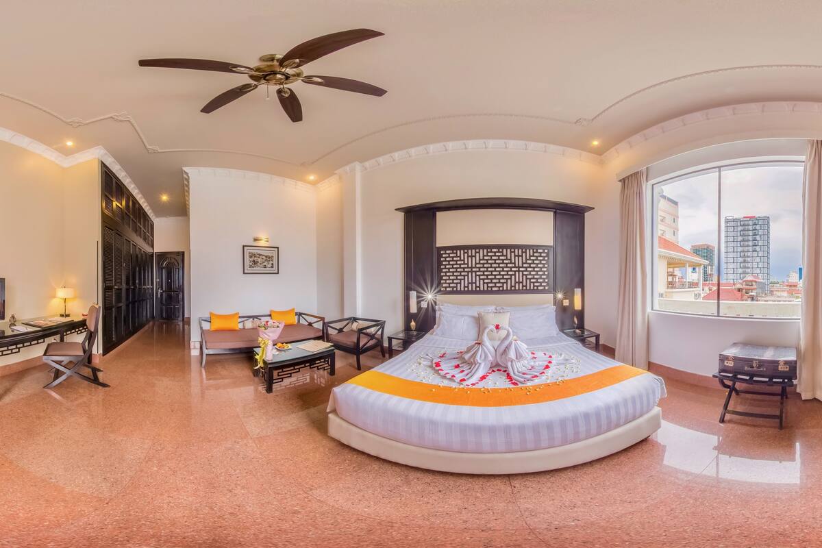 A spacious hotel room with a large bed, a sitting area with a sofa and chairs, a desk with a lamp, and a large window offering a city view.