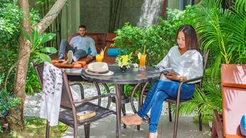 Un área de estar al aire libre con ambiente tropical, con mesas y sillas metálicas, rodeada de exuberante vegetación y un muro de piedra.