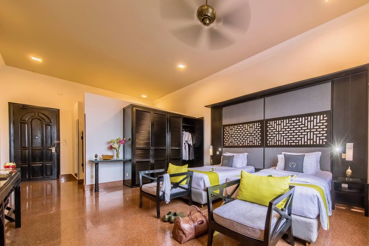 A hotel room with two beds, a desk, a chair, a wardrobe, and a ceiling fan.