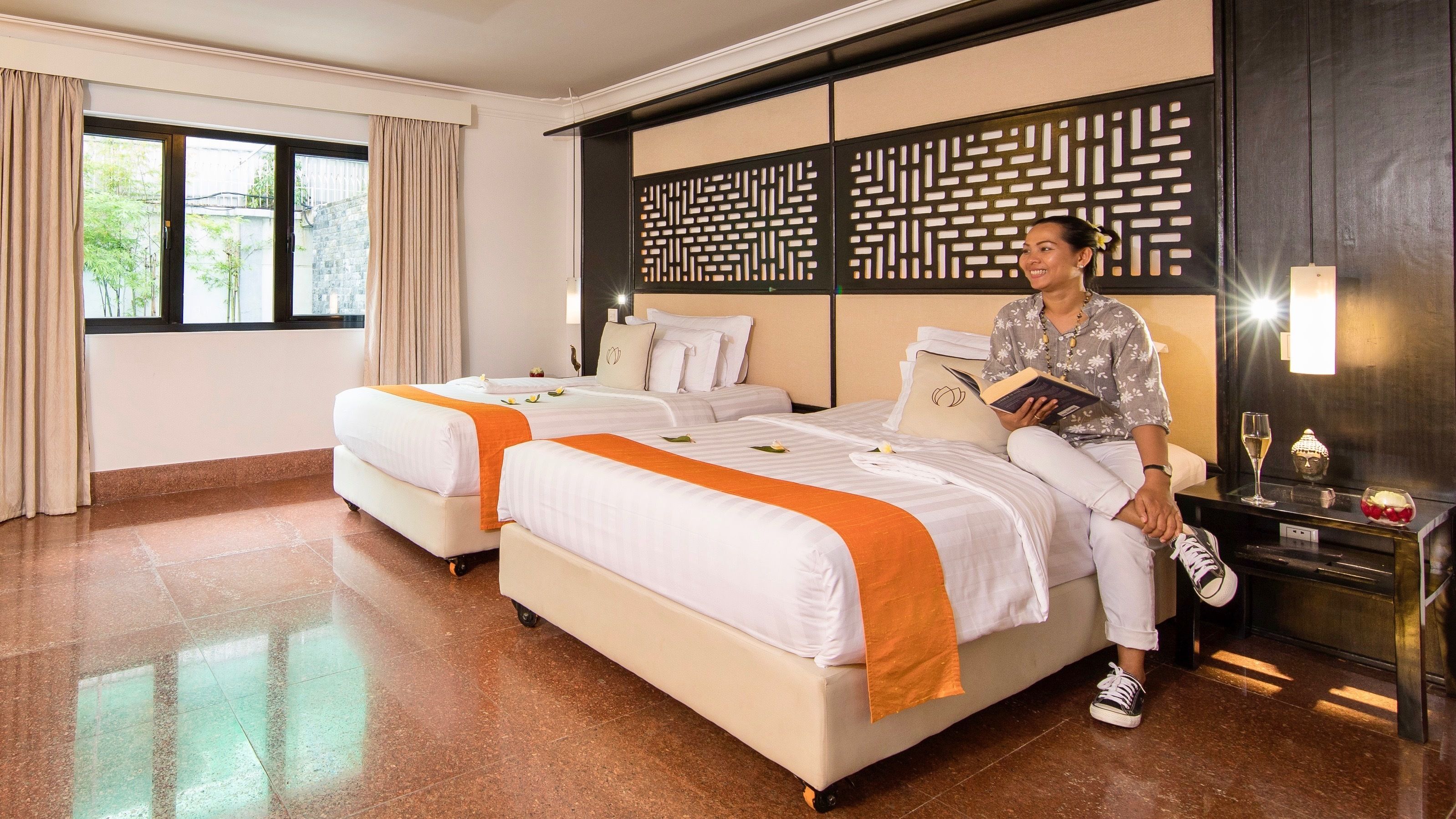 A hotel room with two beds, a large window, a decorative wall panel, and a bedside table with a book and a glass.