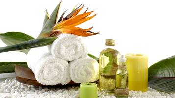 A spa setting with a rolled white towel, a green candle, a bottle of oil, and a yellow candle on a bed of white pebbles.