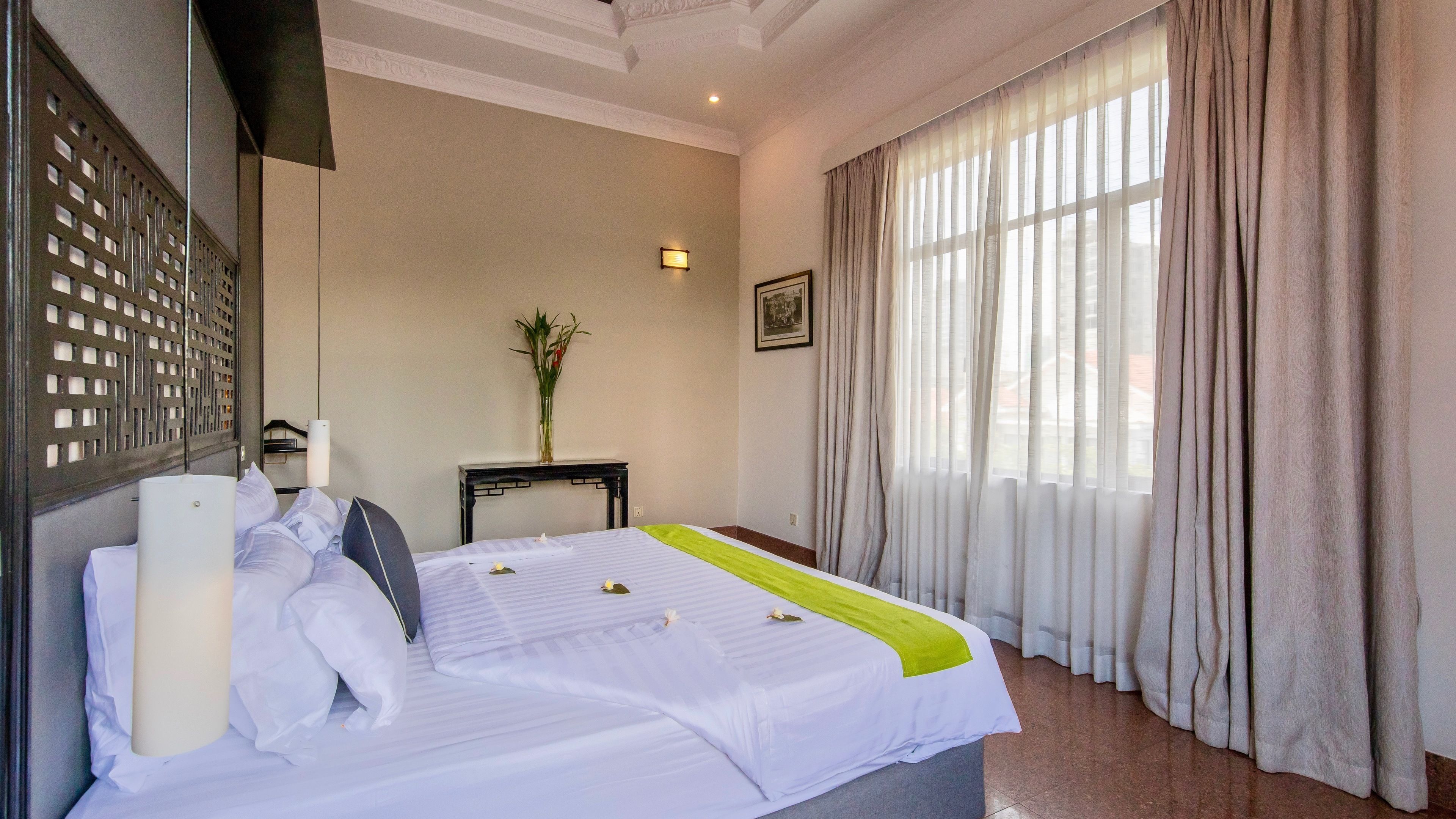 A hotel room with a neatly made bed, a small table with a vase, and a window with curtains.