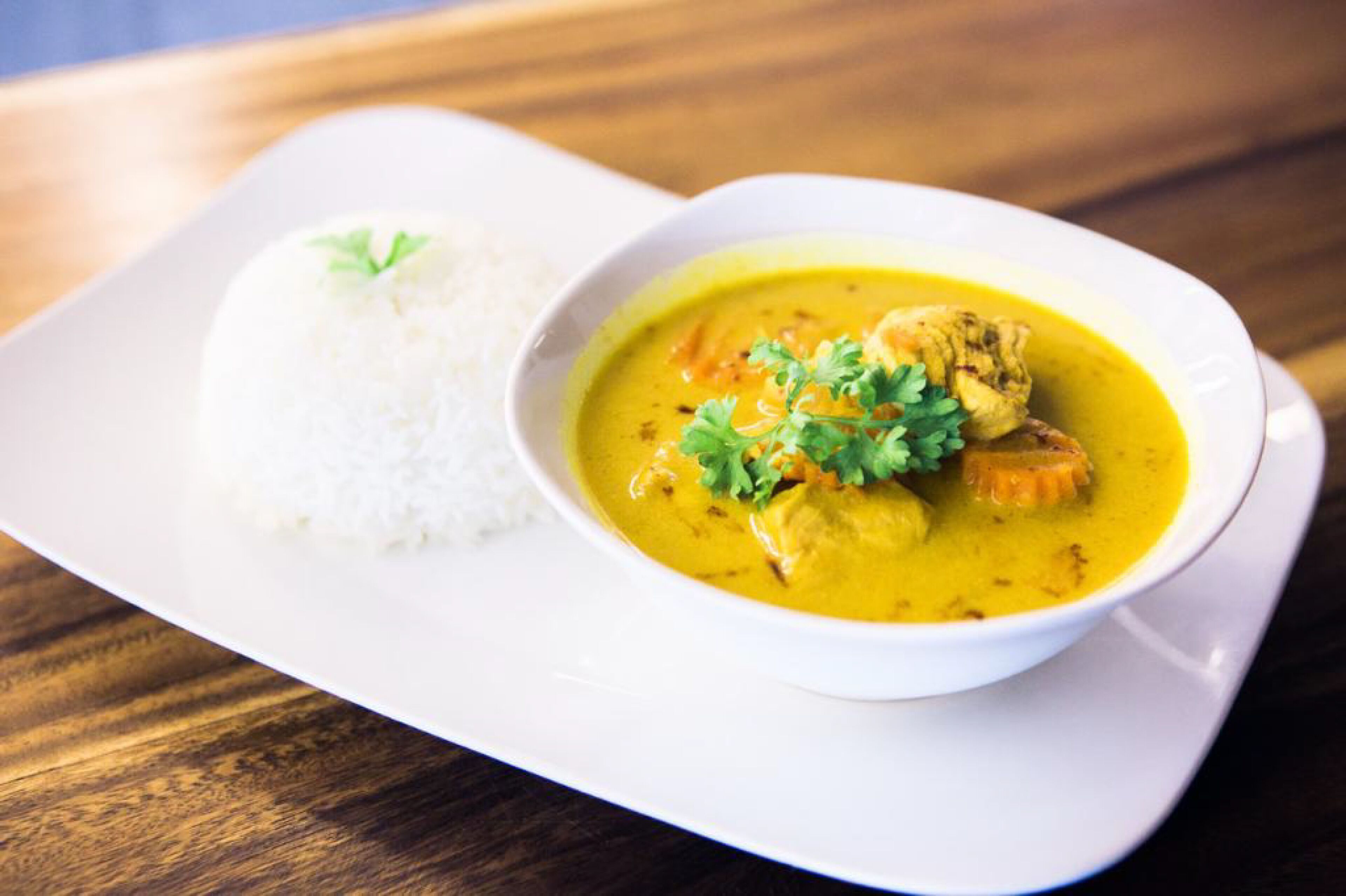Een witte kom met curry, kip en rijst op een wit bord.