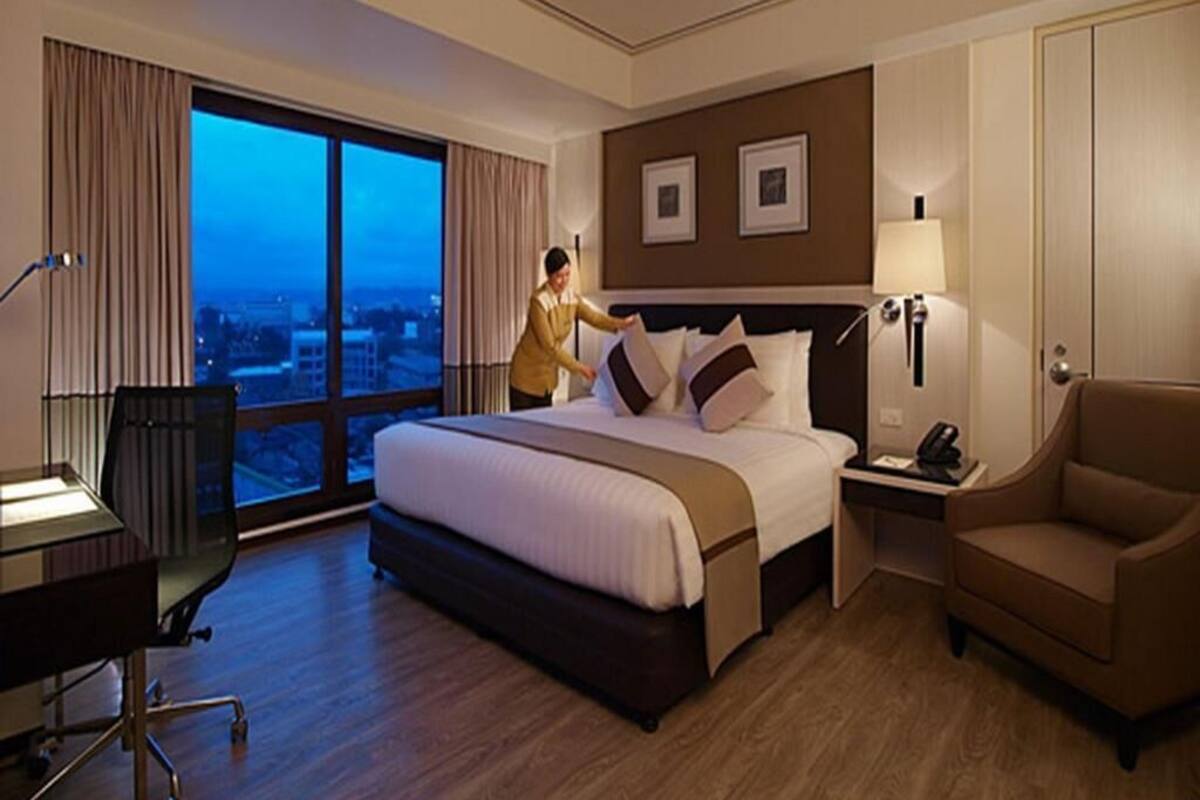 A hotel room with a large bed, a desk, a chair, a nightstand, and a view of the city through large windows.