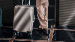 Eine Person in einem beigen Anzug trägt einen Koffer in einem Aufzug mit metallischen Wänden und einem Bedienfeld.