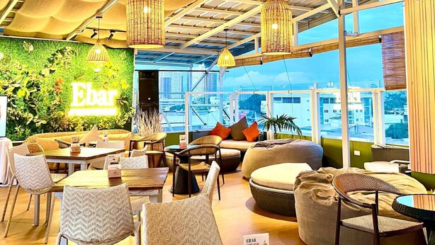 A modern lounge area with wicker furniture, a green wall with 'Ebar' signage, and a view of the cityscape through large windows. - The Upperhouse Hotel – Boutique Stay with City & River Views (Cagayan de Oro)