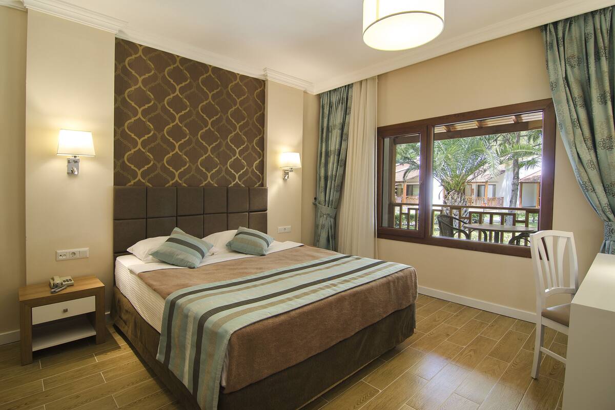 Una camera d'albergo con un letto, comodino, lampada, finestra con tende e vista all'esterno.