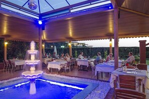 An outdoor dining area with tables and chairs, a fountain, and a pool. - Kustur Club Holiday Village (Kusadasi)