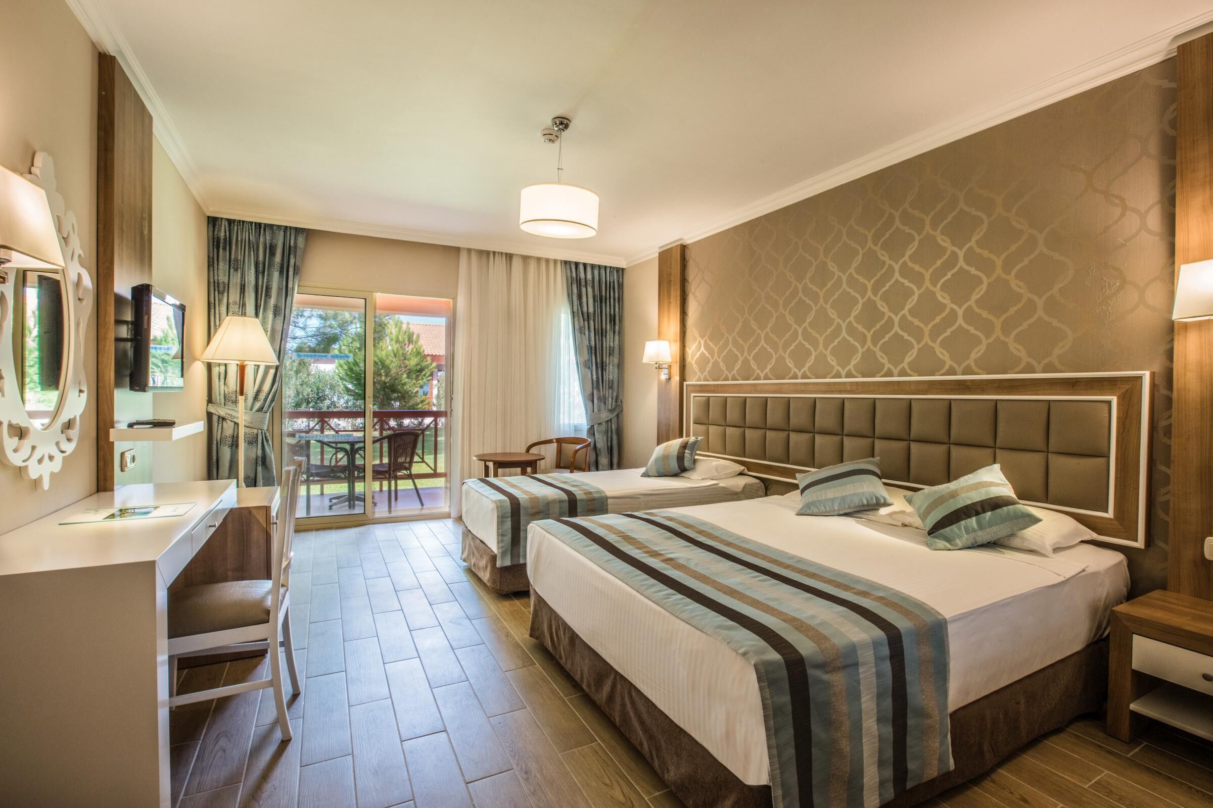A hotel room with two beds, a desk, a chair, a TV, and a balcony with a table and chairs.