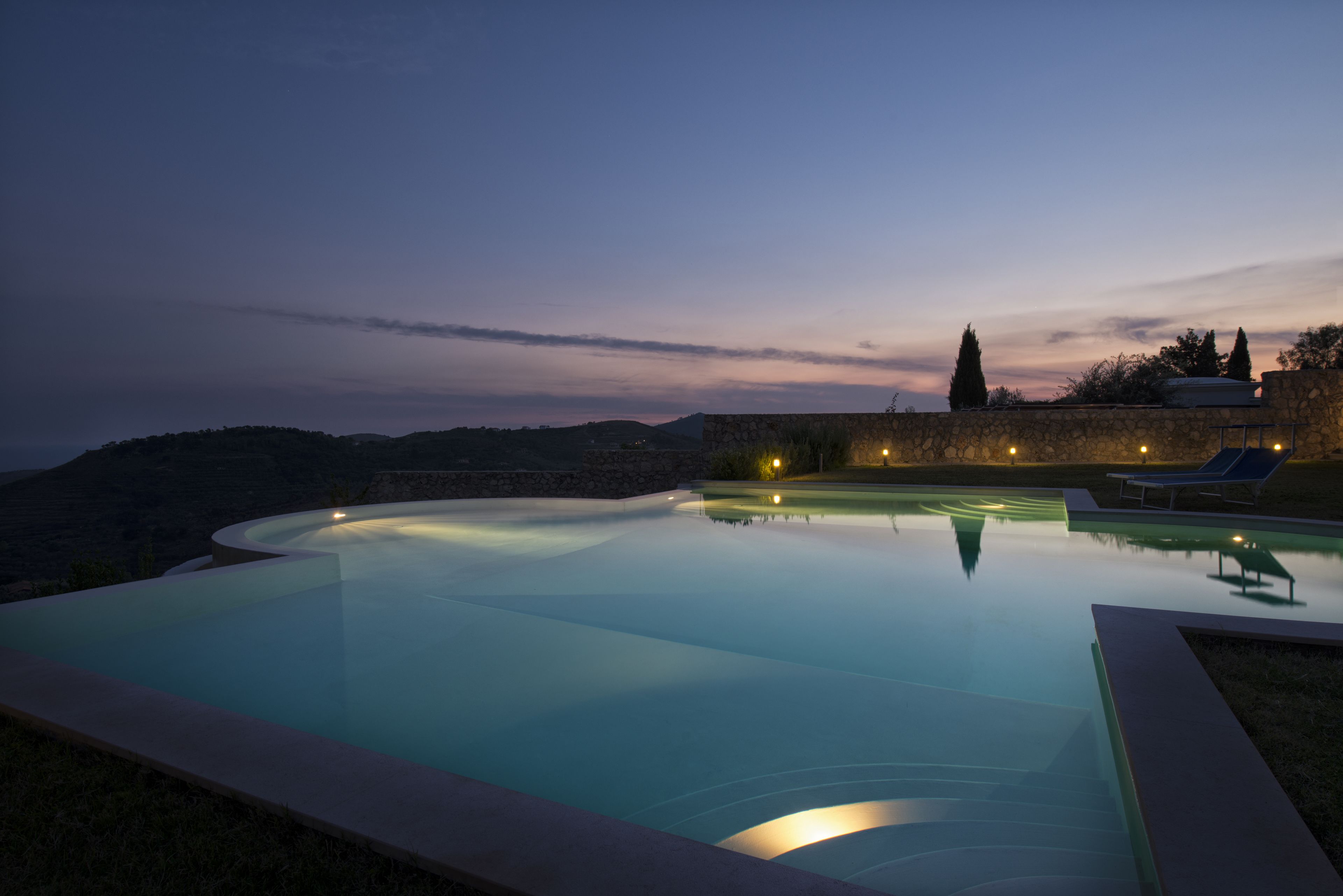 A serene infinity pool at dusk with a stone wall and lounge chairs.