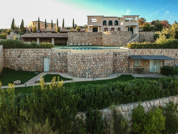 A large stone wall with a swimming pool, a patio area, and a view of surrounding buildings.
