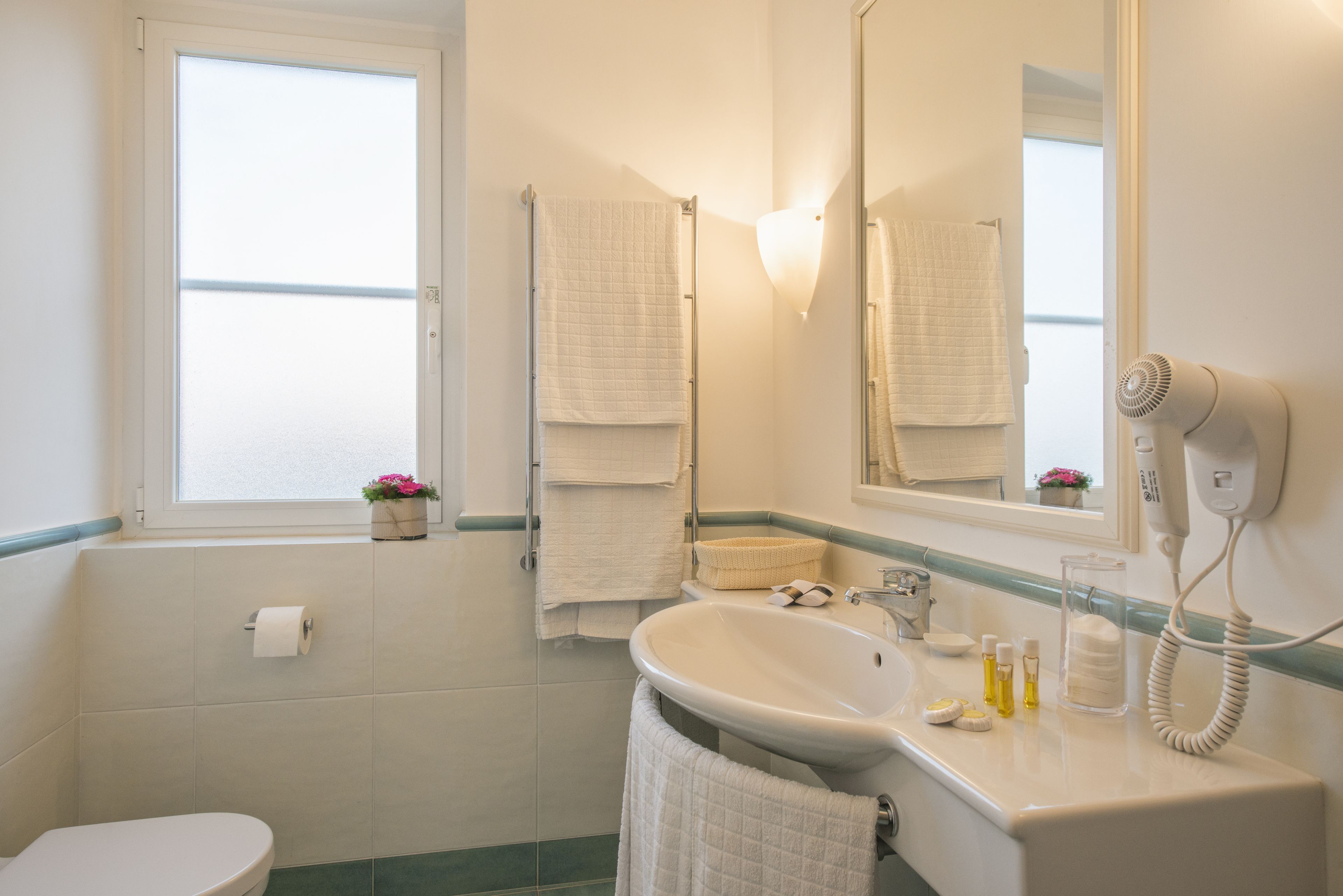 Un bagno con il water, un lavandino e uno specchio. C'è un portasciugamani con diversi asciugamani, un asciugacapelli e una finestra con vista.