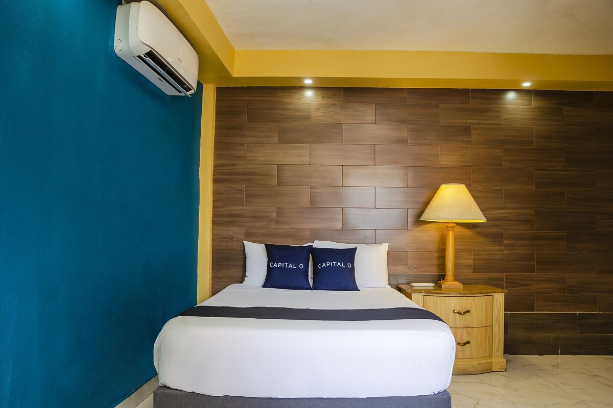 A hotel room with a bed, a nightstand, a lamp, and a wall-mounted air conditioner.