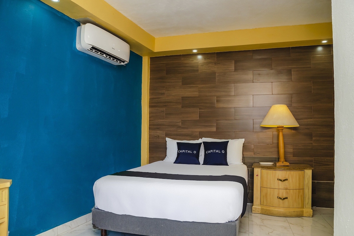 A hotel room with a bed, a nightstand, a lamp, an air conditioning unit, and a wooden headboard.