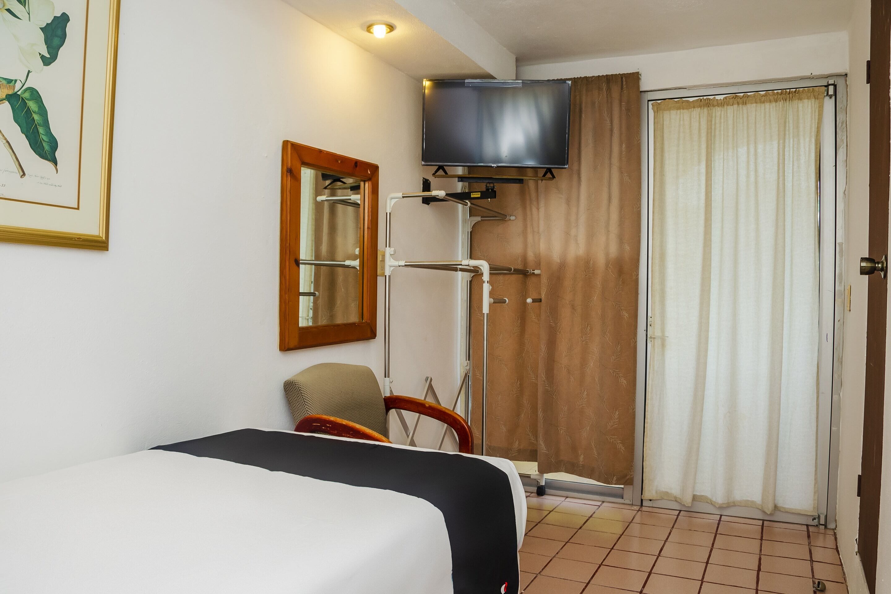 Quarto de hotel com cama, poltrona, televisão fixada na parede, espelho e porta coberta por cortina.