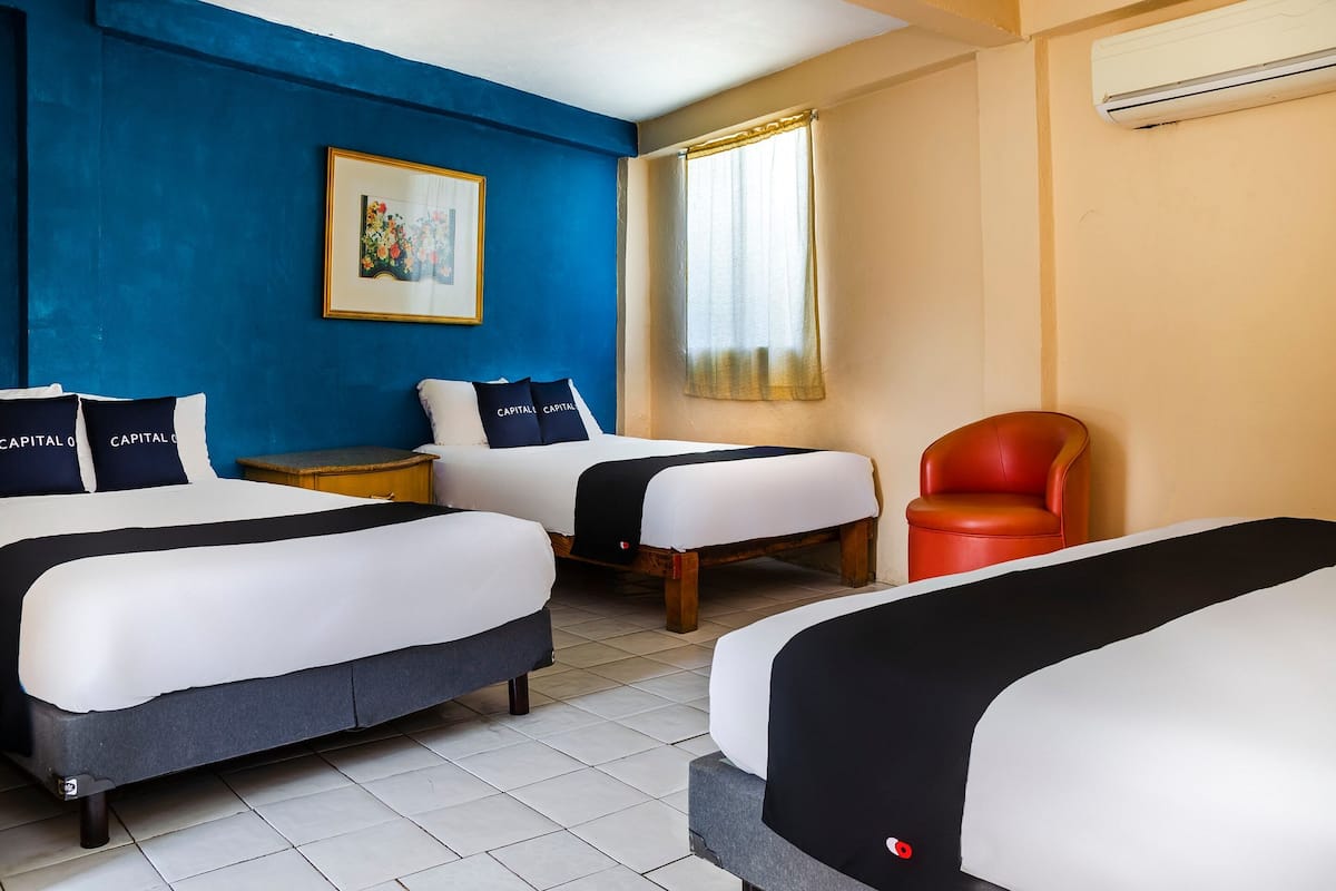 A hotel room with three beds, a red chair, a framed picture on the wall, and an air conditioning unit.