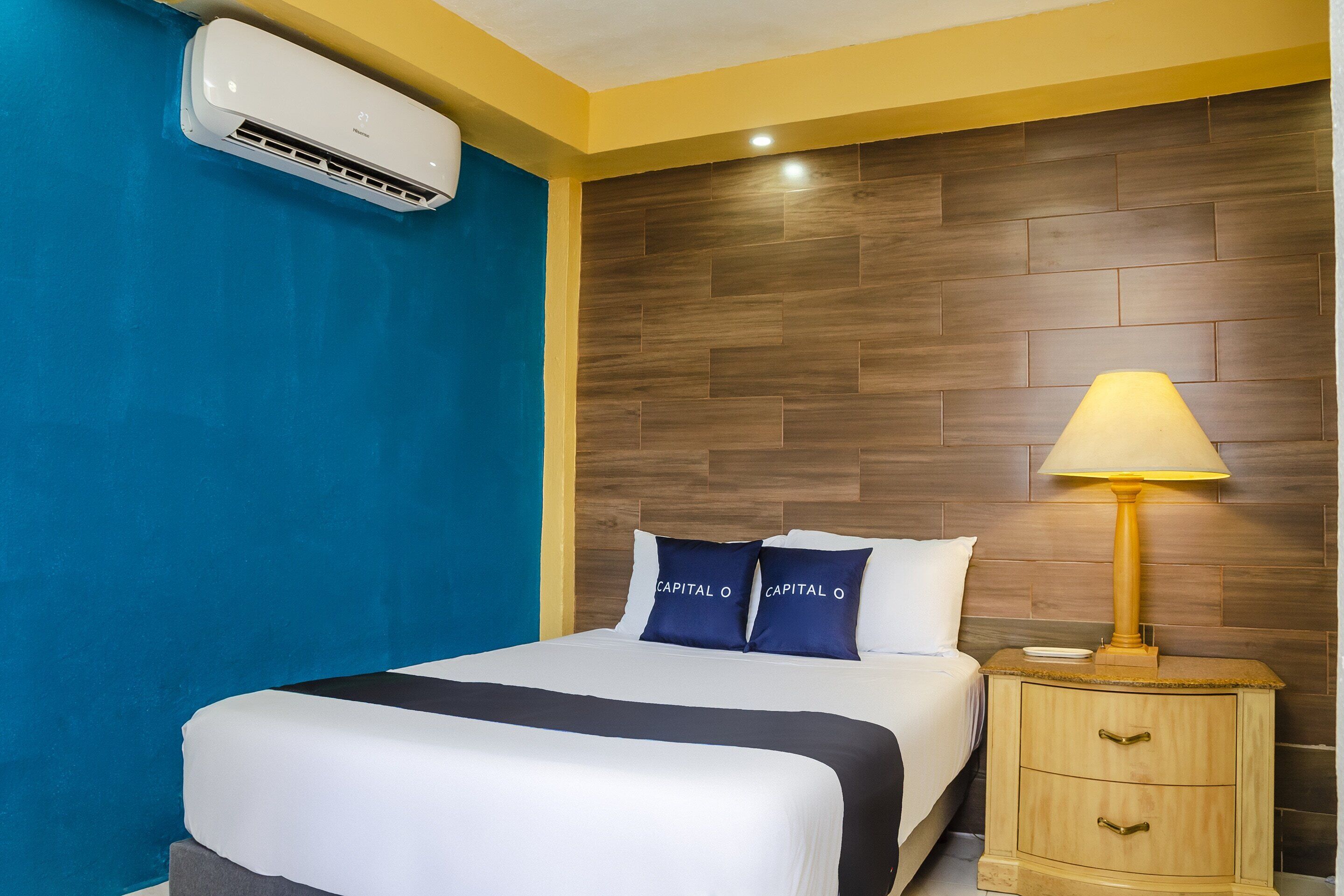Een hotelkamer met een bed, nachtkastje, lamp en airconditioning.