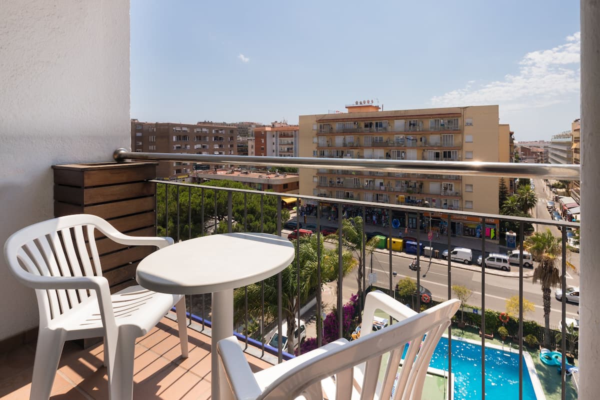 Un balcon avec une table et des chaises, donnant sur une rue bordée d’immeubles et sur une piscine.