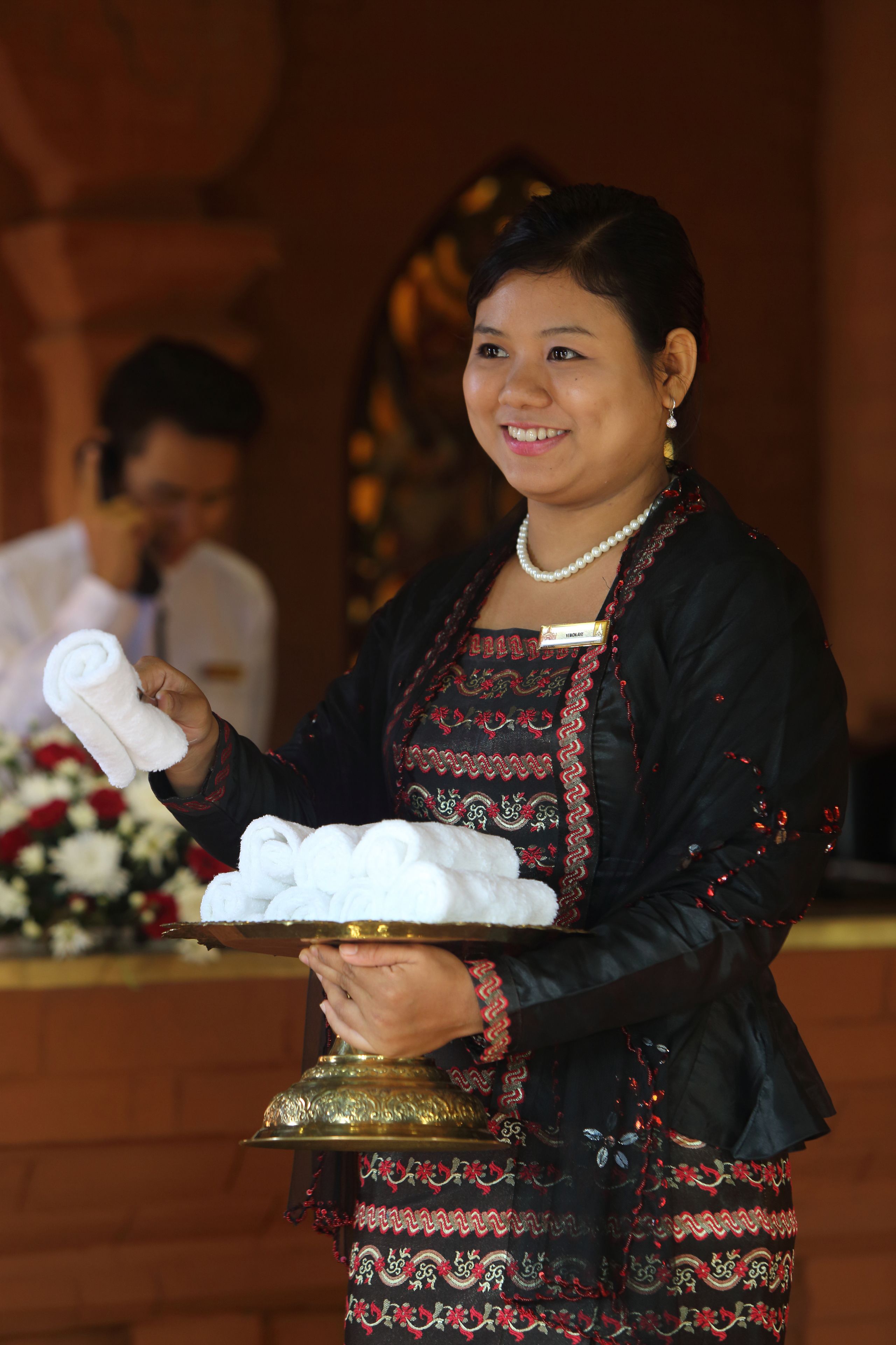 A person holding a tray with rolled towels, standing in front of a counter with a floral arrangement.