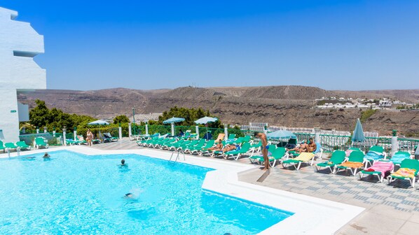 A pool area with multiple sun loungers and umbrellas, surrounded by a white building and a view of distant hills. - Apartamentos Monteparaiso (Mogan)