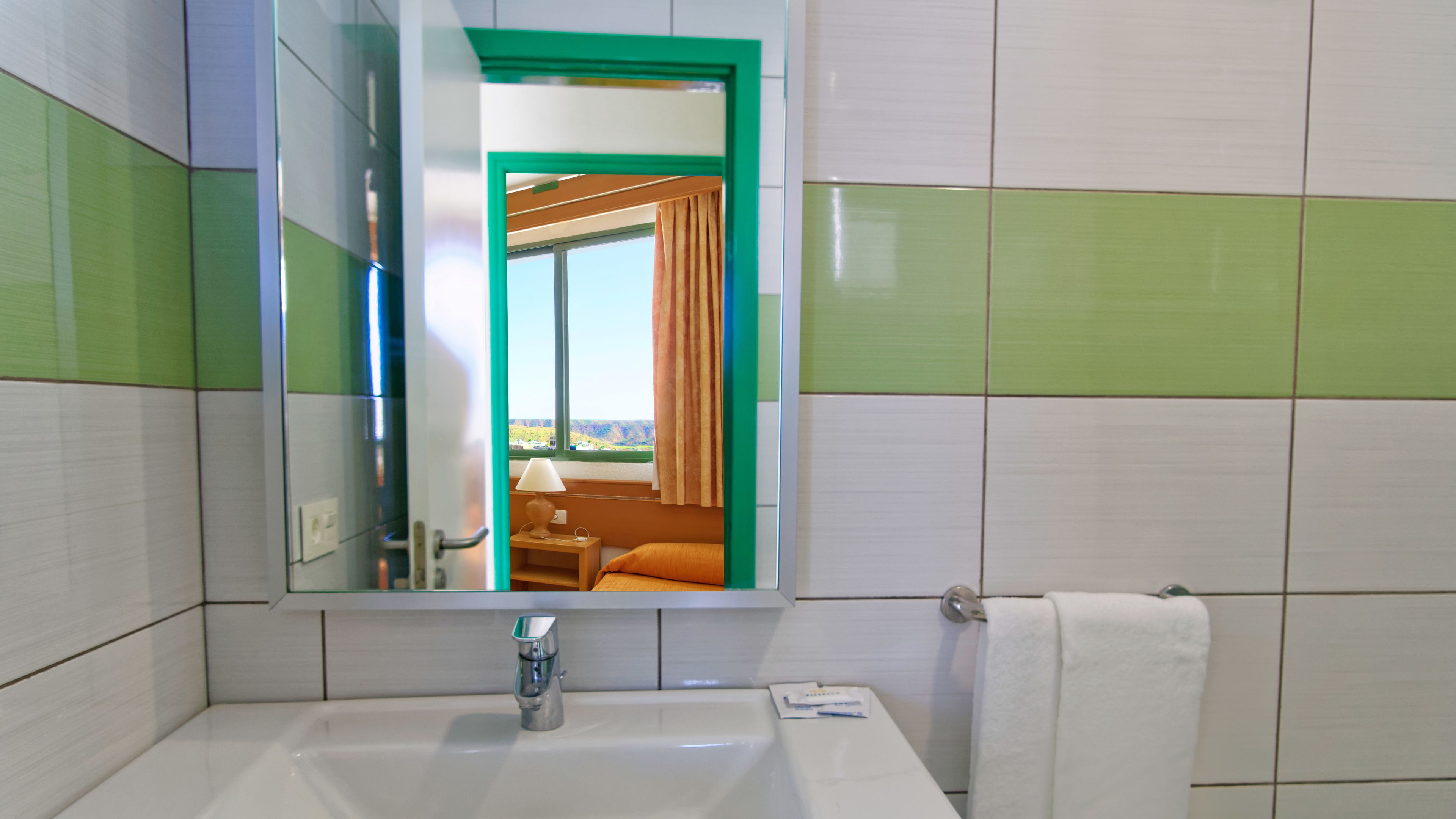 Une salle de bain avec un lavabo, un miroir et un porte-serviettes. Une fenêtre donne sur une pièce où se trouvent un lit et des rideaux.
