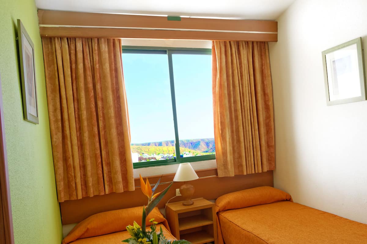 A hotel room with two beds, a window with curtains, a nightstand with a lamp, and a view of a landscape.