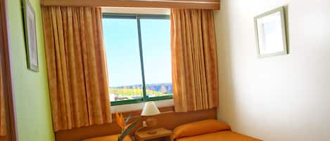 A hotel room with two beds, a window with curtains, a nightstand with a lamp, and a view of a landscape.