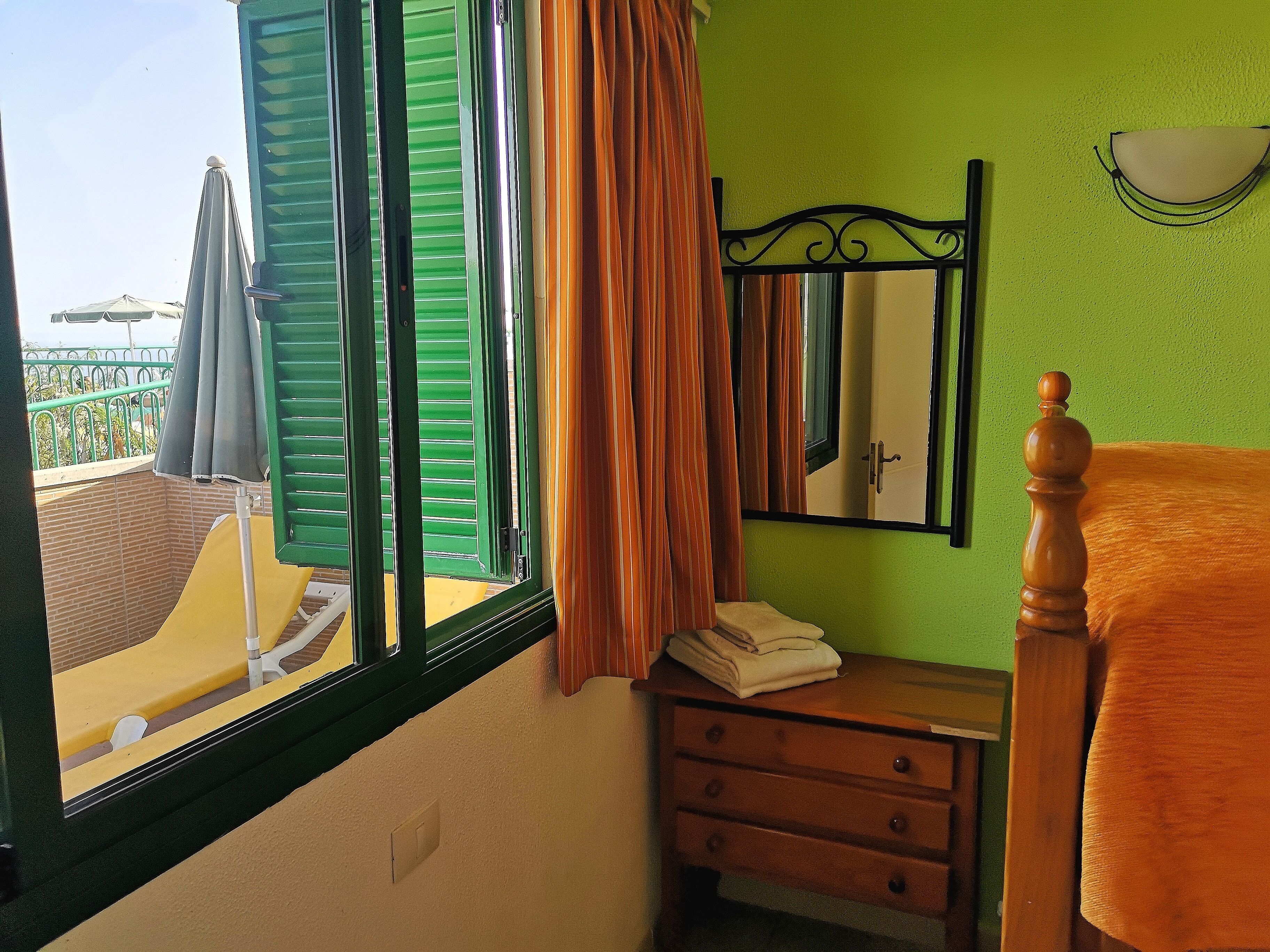 A room with a bed, a nightstand, a mirror, and a window with green shutters.