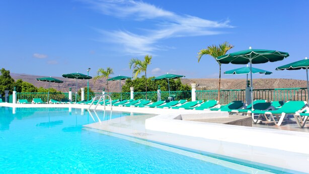 A pool area with green lounge chairs and umbrellas, a clear blue sky, and a mountainous backdrop. - Apartamentos Monteparaiso (Mogan)