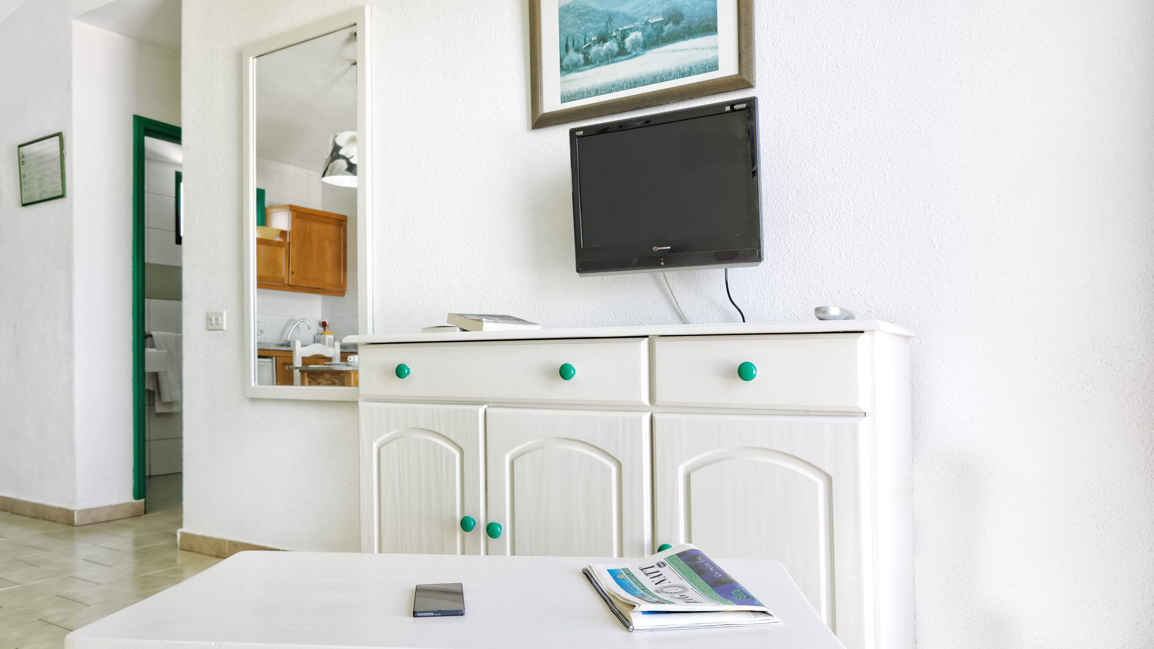 Una habitación blanca con un televisor de pantalla plana sobre un mueble blanco con pomos verdes, una mesa con un libro y un teléfono, y un espejo que refleja una cocina.