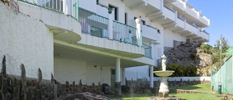 A multi-story building with balconies, a stone wall, and a fountain.