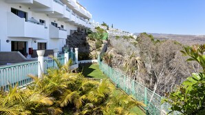 A white building with balconies, a green fence, and a view of a hillside with sparse vegetation. - Apartamentos Monteparaiso (Mogan)